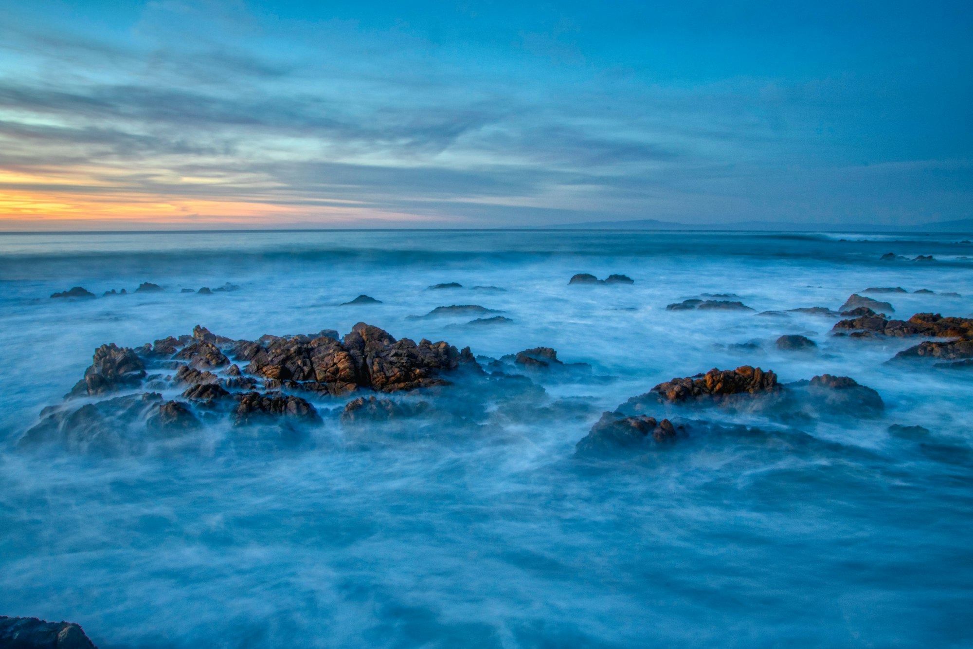 Long-exposure photo of waves washing over rocks along the Asilomar coast at dusk, with a band of sunset light on the horizon.