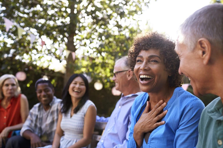 A group of adults sitting together outdoors under trees, laughing; a woman in a blue shirt in the foreground has her hand on her chest mid-laugh.